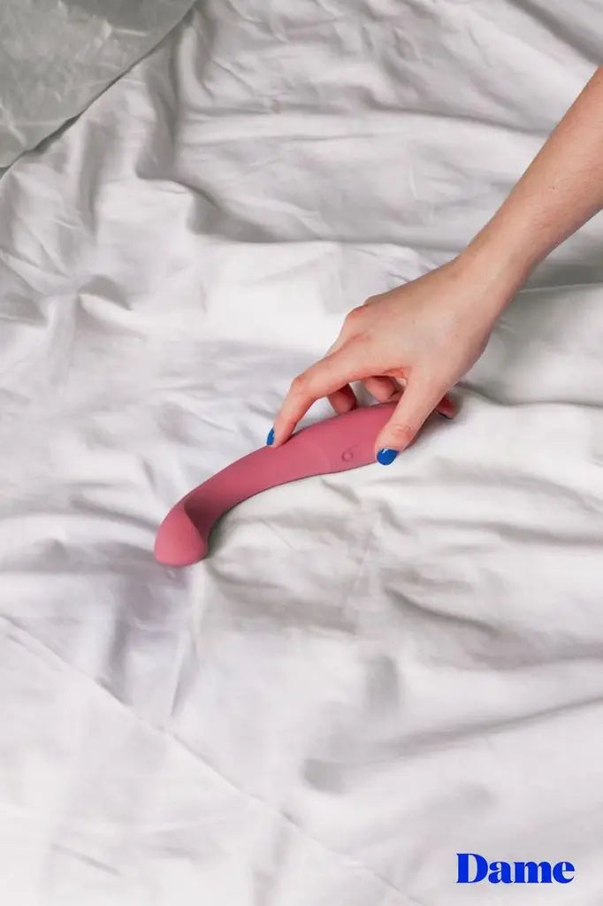 A person with blue nail polish holds a pink arc, g-spot vibrator against white sheets